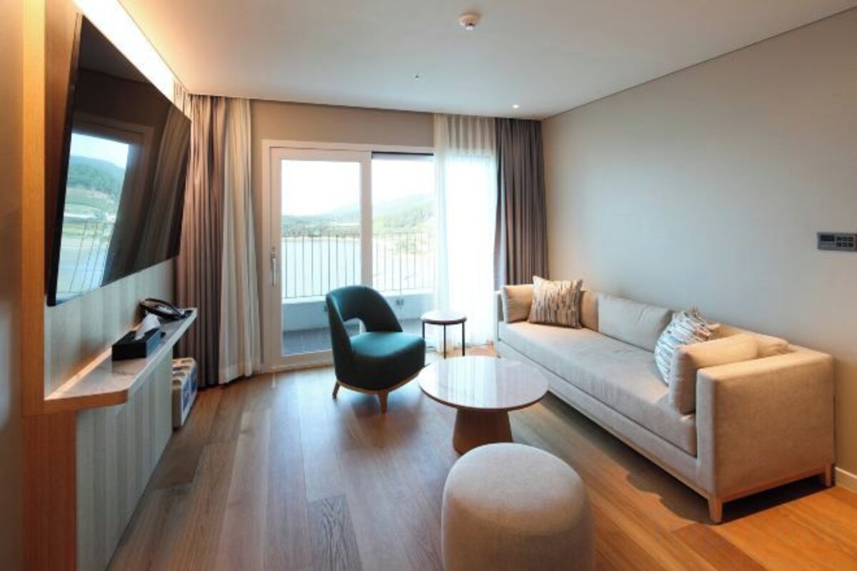 A modern hotel room with a sofa, armchair, round coffee table, and a TV mounted on the wall.
