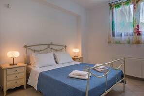 A neatly made bed with a blue bedspread, two bedside lamps, and a window with floral curtains. - Villa Katerina (Chania)