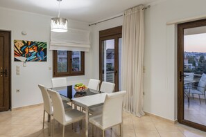 A dining area with a table, chairs, and a fruit bowl. There is a window with a view of buildings and a door to the left. - Villa Katerina (Chania)