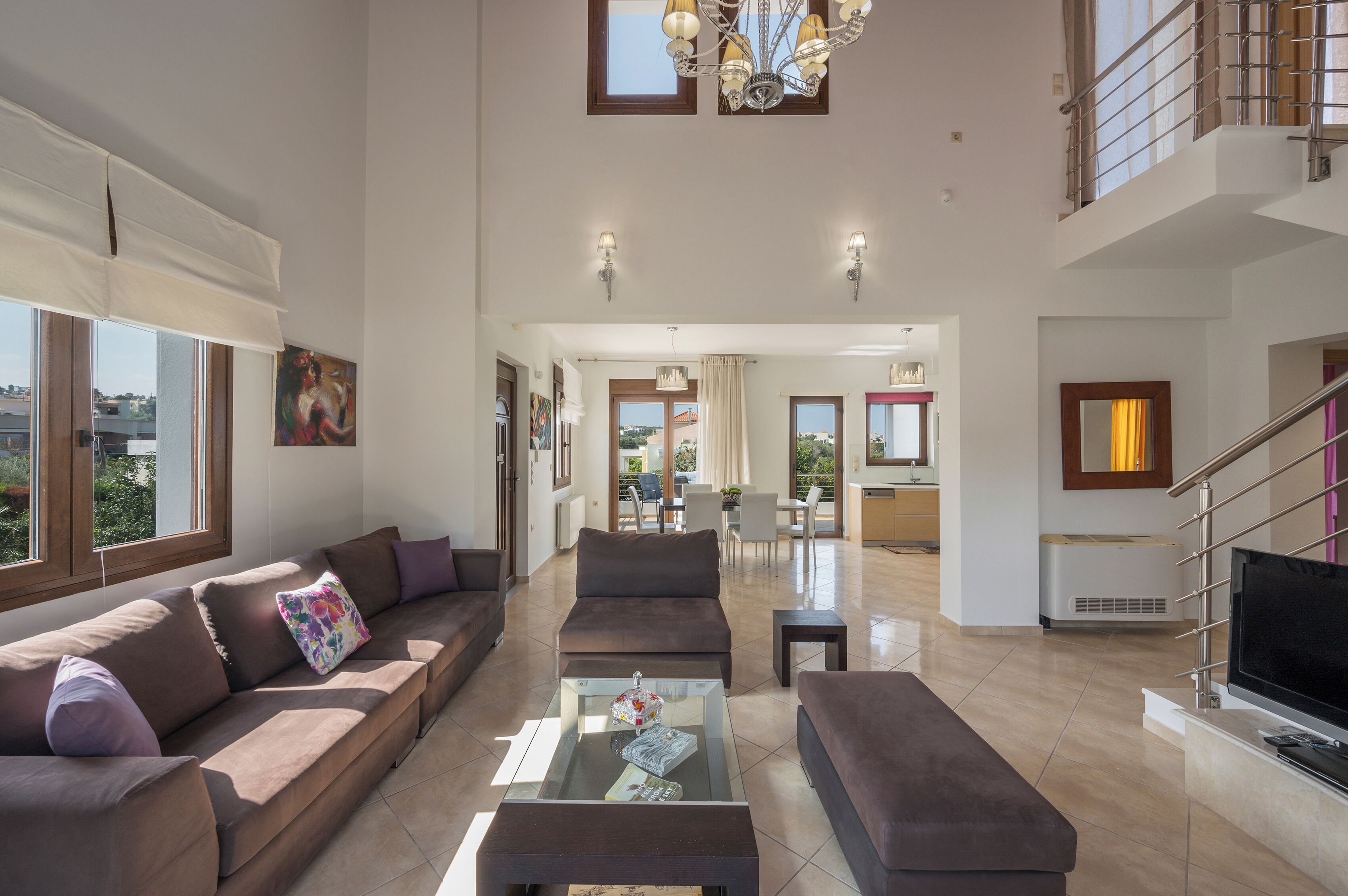 A spacious living area with a large sofa, a coffee table, and a television. There is a staircase leading to an upper level, and a dining area is visible in the background.