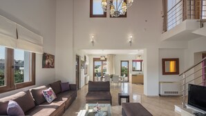 A spacious living area with a large sofa, a coffee table, and a television. There is a staircase leading to an upper level, and a dining area is visible in the background. - Villa Katerina (Chania)