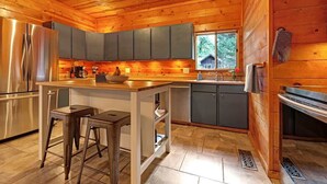 A modern kitchen with wooden walls, a stainless steel refrigerator, and a large island with stools. - Charming 3BR with Hot Tub, Balcony & Wood Stove (Guerneville)