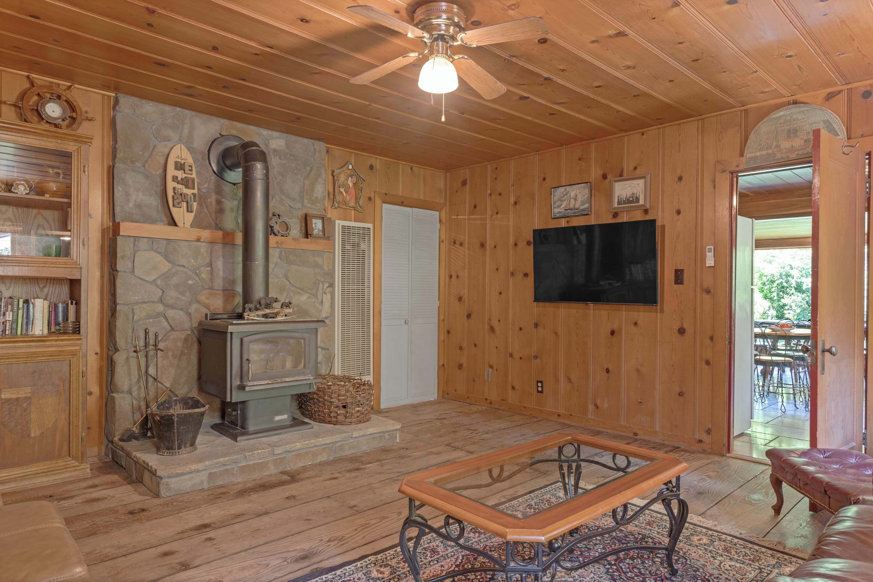 A cozy cabin interior with a stone fireplace, wooden walls, a ceiling fan, and a television mounted on the wall.