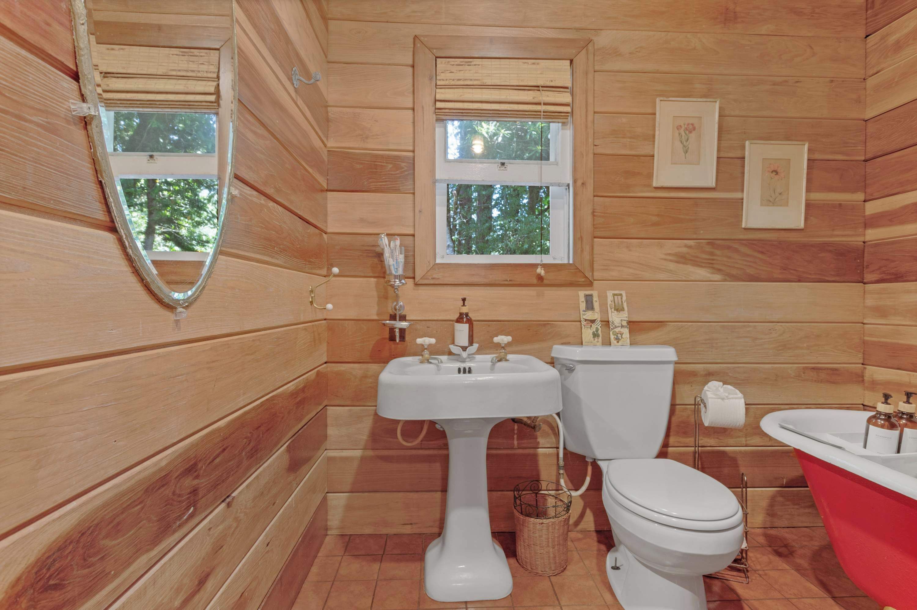 A wooden-paneled bathroom with a pedestal sink, toilet, and red freestanding bathtub.
