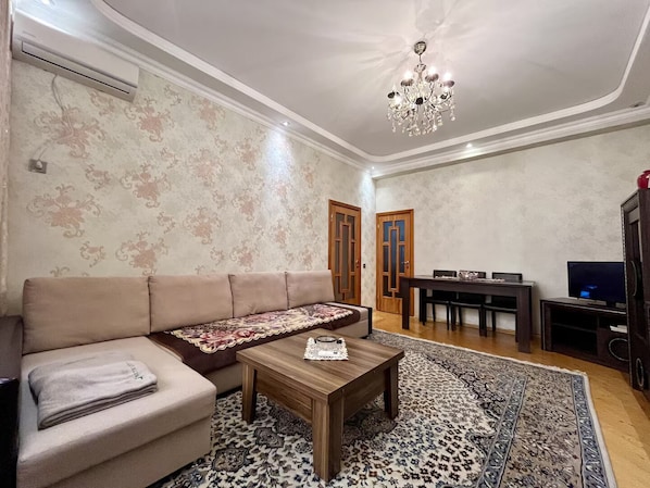 A living room with a sofa, a coffee table, a chandelier, a TV, and a patterned rug.