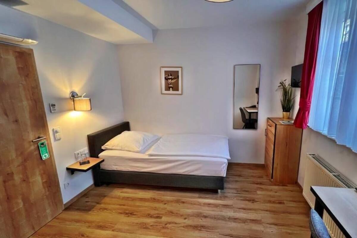 Un dormitorio con cama, mesita de noche, lámpara, espejo y ventana con cortinas.