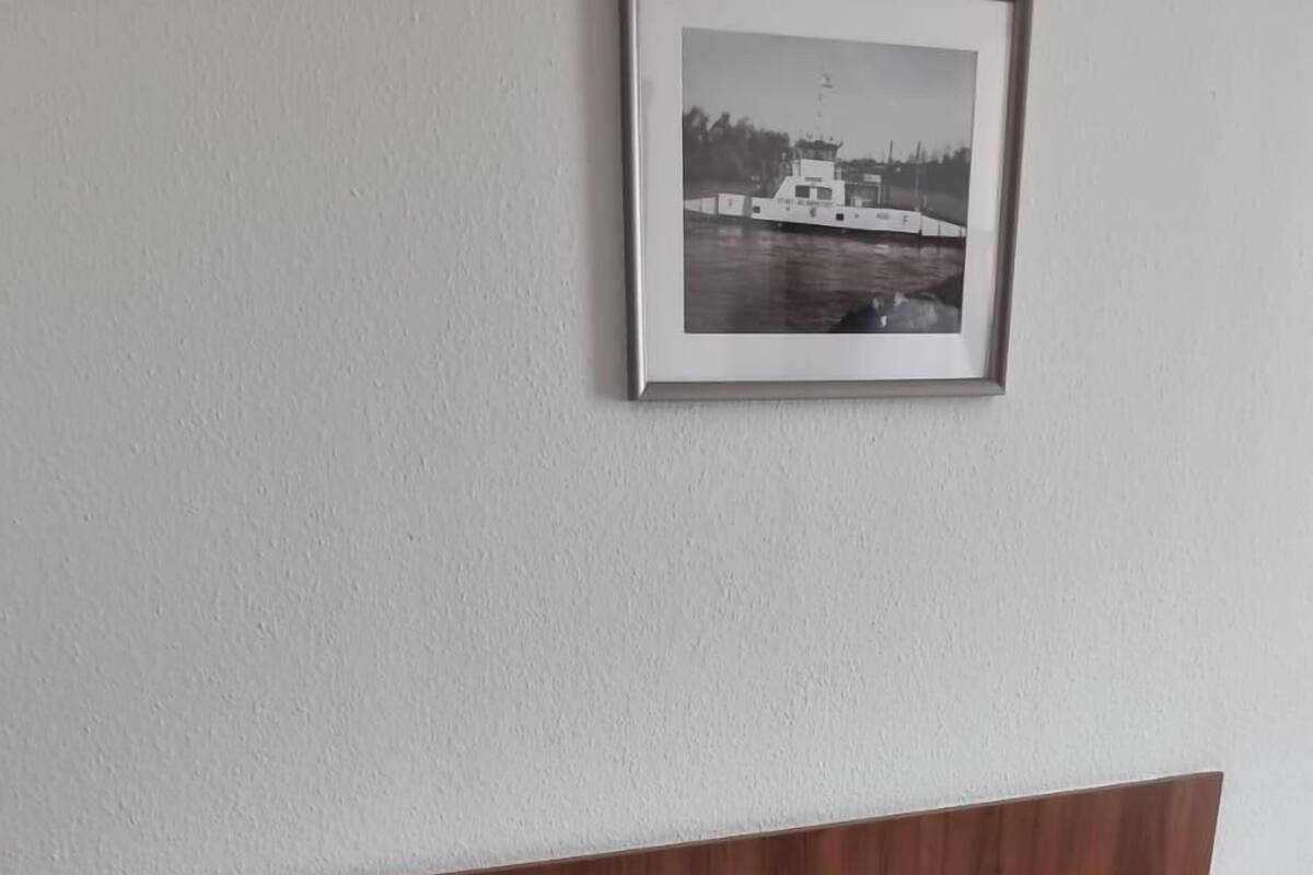 Una cama bien hecha con sábanas blancas, cabecera de madera, mesita de noche con lámpara y un cuadro enmarcado en la pared.