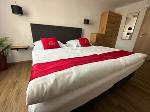 En pænt redt seng med hvidt og rødt sengetøj, et lille natbord med en lampe, og en træskab i et moderne rum.