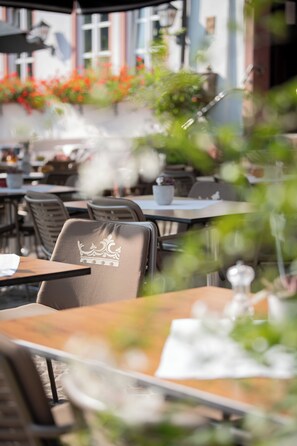 En restaurant med udendørs siddeplads, herunder borde og stole, samt et dekorativt krone-emblem på en stol.
