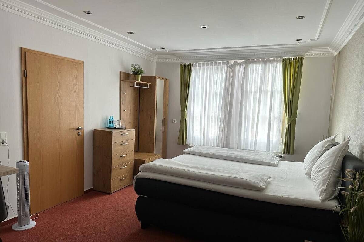 Un dormitorio con cama, armario, espejo, mesita de noche y ventana con cortinas.
