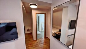 A narrow hallway with a wooden floor leading to a balcony with a glass door. A flat-screen TV is mounted on the wall to the left, and a mirror is on the right. - Zu den drei Kronen (Seligenstadt)