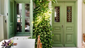 Une porte verte avec une fenêtre, une table blanche ornée d’un vase de fleurs et un mur vert recouvert de plantes grimpantes.