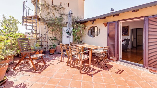 A patio with wooden furniture and potted plants, a spiral staircase, and a building with a round window and wooden shutters. - Colosseum Terrace Penthouse (Rome)