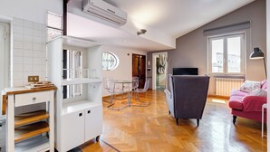A modern living room with a wooden herringbone floor, a grey armchair, a pink sofa, and a glass dining table with chairs. - Colosseum Terrace Penthouse (Rome)