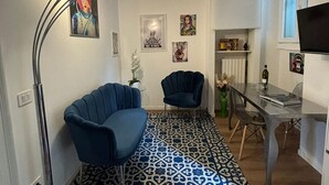 Een moderne woonkamer met een gestreept vloerkleed, twee blauwe fauteuils, een glazen eettafel en een televisie aan de muur.