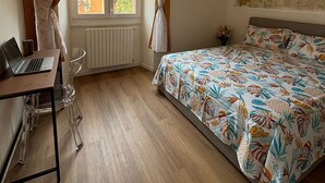 Een slaapkamer met een houten vloer, een bed met een bloemenprint, een bureau met een laptop en een raam met gordijnen.