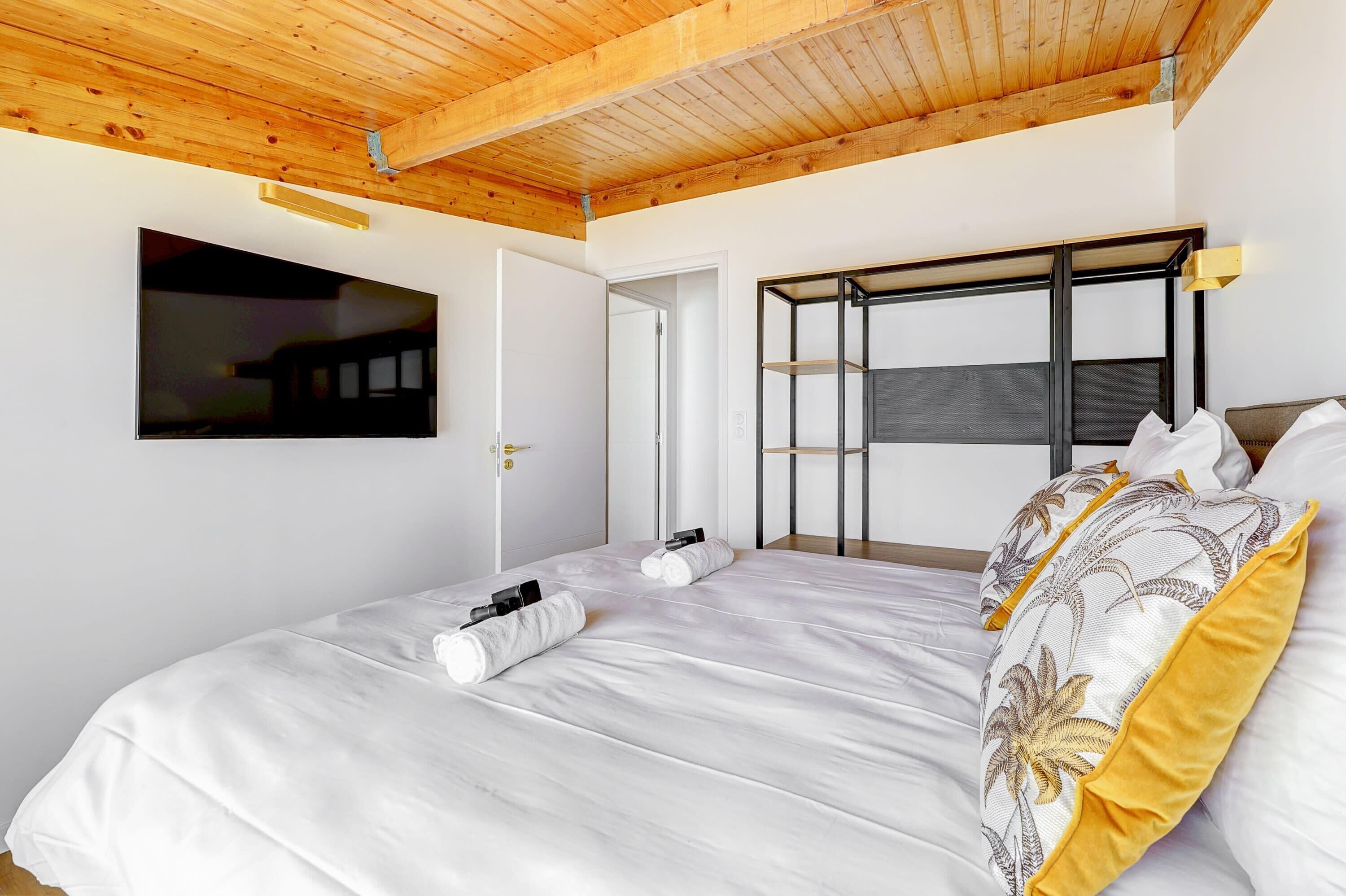 Une chambre avec un plafond en bois, un lit avec une literie blanche, un téléviseur fixé au mur et une porte donnant sur une autre pièce.