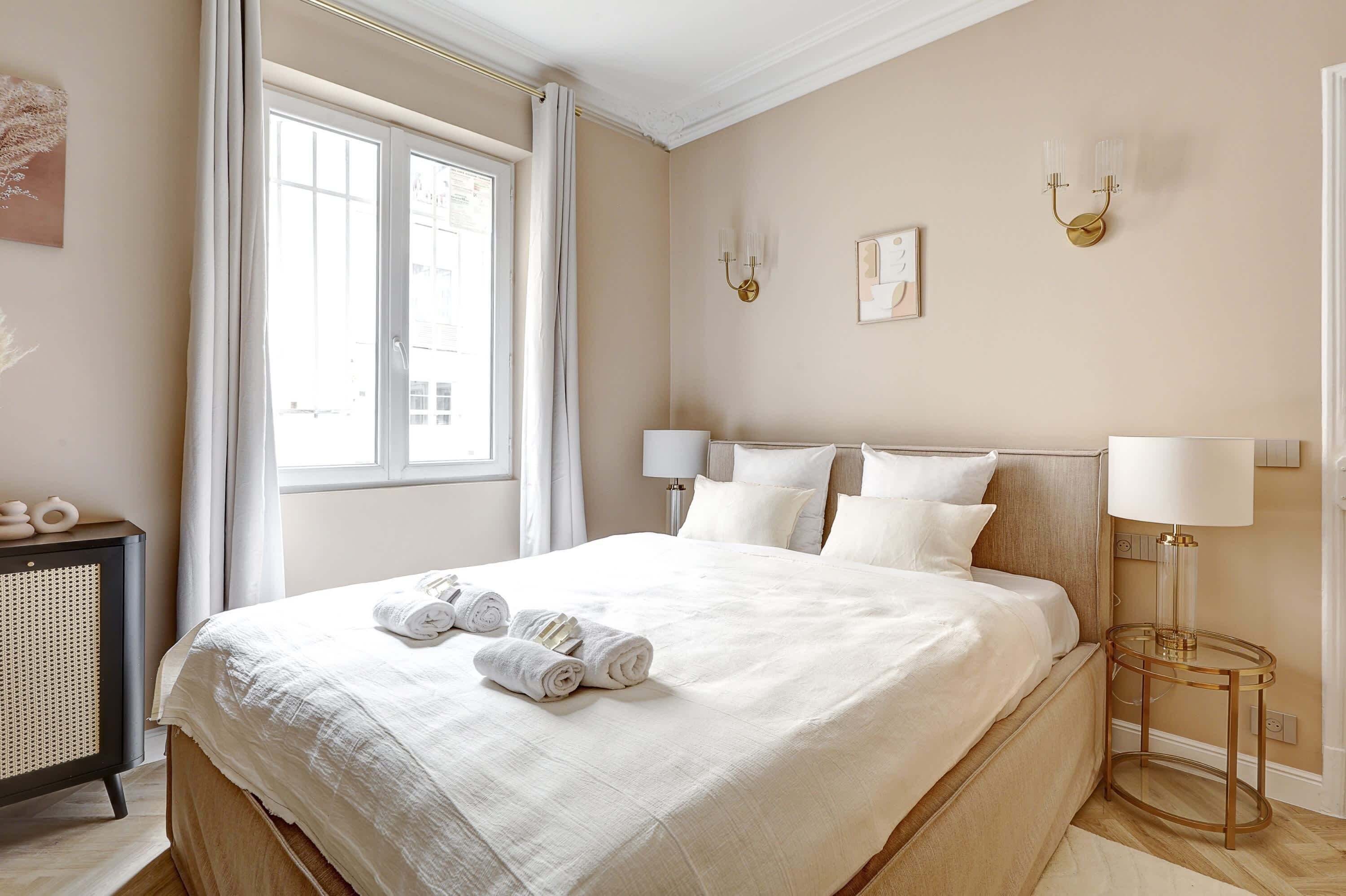 Een net opgemaakt bed met witte lakens en opgerolde handdoeken, een bedlampje en een wandlamp.