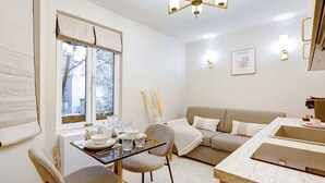 A modern living room with a sofa, a dining table with chairs, a kitchen area, and a large window with blinds.
