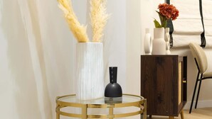A corner of a room with a round glass table with gold legs, a white vase with pampas grass, and a small black vase.