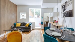 En moderne stue med en sofa, et spisebord dækket til seks personer, og et køkkenområde, der kan ses gennem en åben dør.