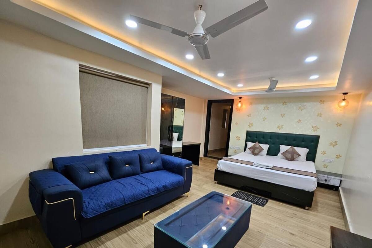A modern hotel room with a blue sofa, a glass coffee table, a bed with a green headboard, and a ceiling fan.