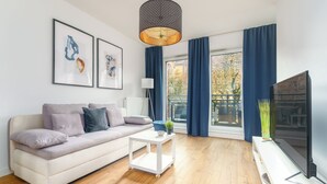 A modern living room with a white sofa, a small white side table, a floor lamp, a large flat-screen TV, and a window with blue curtains. - MARGI Lotus Apartment (Szczecin)