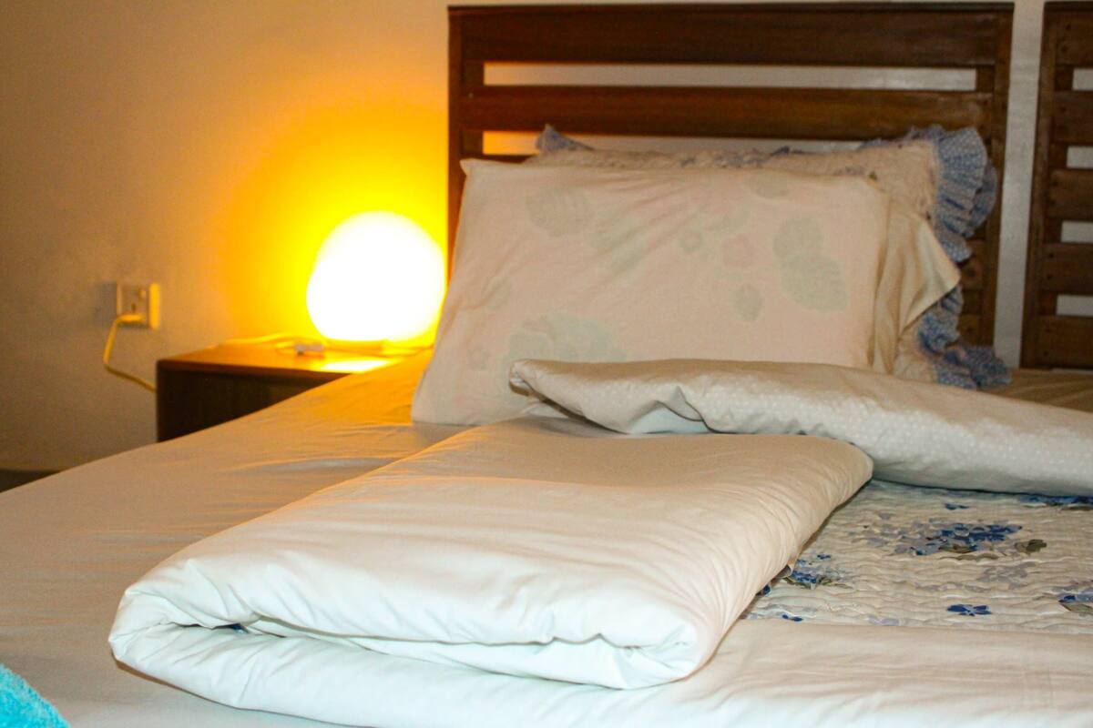 Una cama bien hecha con edredón blanco y almohadas, cabecera de madera y lámpara de noche con luz amarilla.