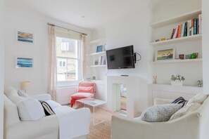 Un salon moderne comprenant un canapé, des fauteuils, une table basse, un téléviseur fixé à un mur blanc, et une bibliothèque remplie de livres et d’objets décoratifs.