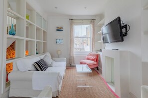 Un salon moderne avec un canapé blanc, un fauteuil rose, une table basse en verre et un téléviseur fixé au mur.