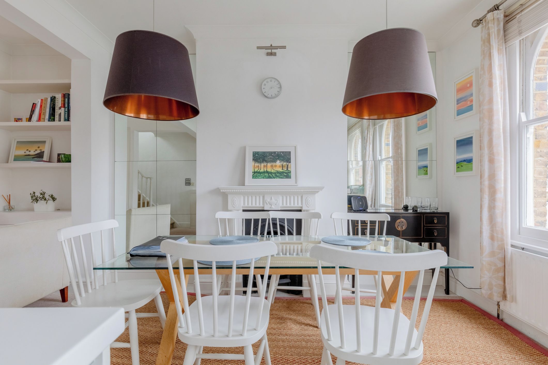 Een eetgedeelte met witte stoelen, een glazen tafel en twee grote hanglampen.