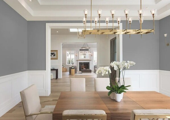 A dining area with a wooden table, white upholstered chairs, a chandelier, and white orchids. - Mandeville Canyon - Exquisite Contemporary Farmhouse with Pool & Spa (Los Angeles)