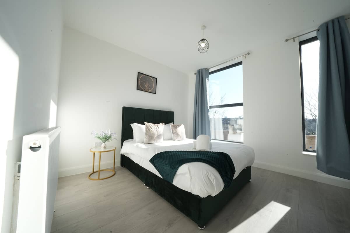 A bedroom with a bed, a small side table, a window with curtains, and a radiator.