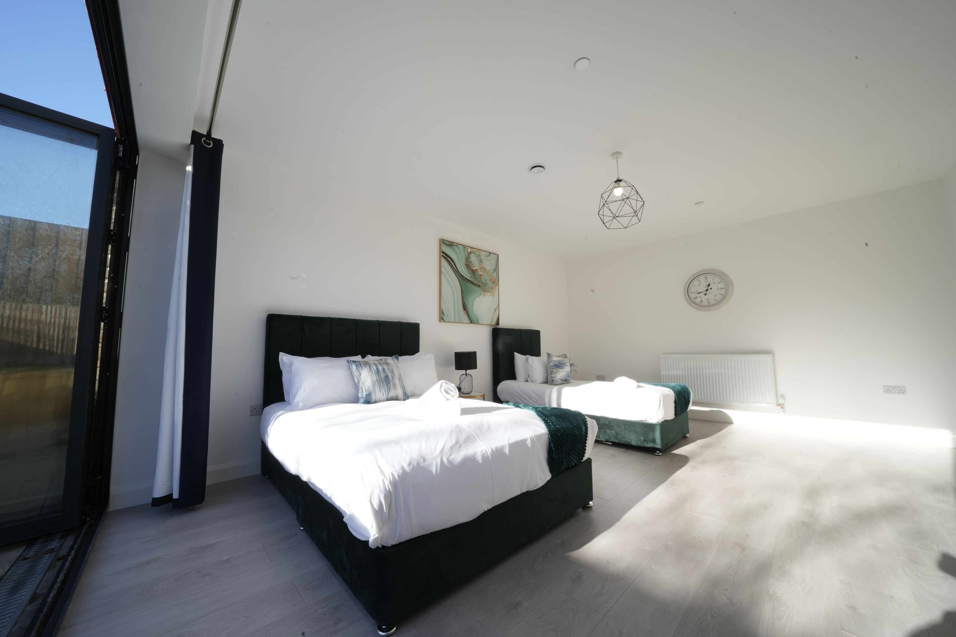 Ein modernes Schlafzimmer mit einem großen Bett, einem kleineren Bett, einem Nachttisch, einer Uhr und einem Fenster.