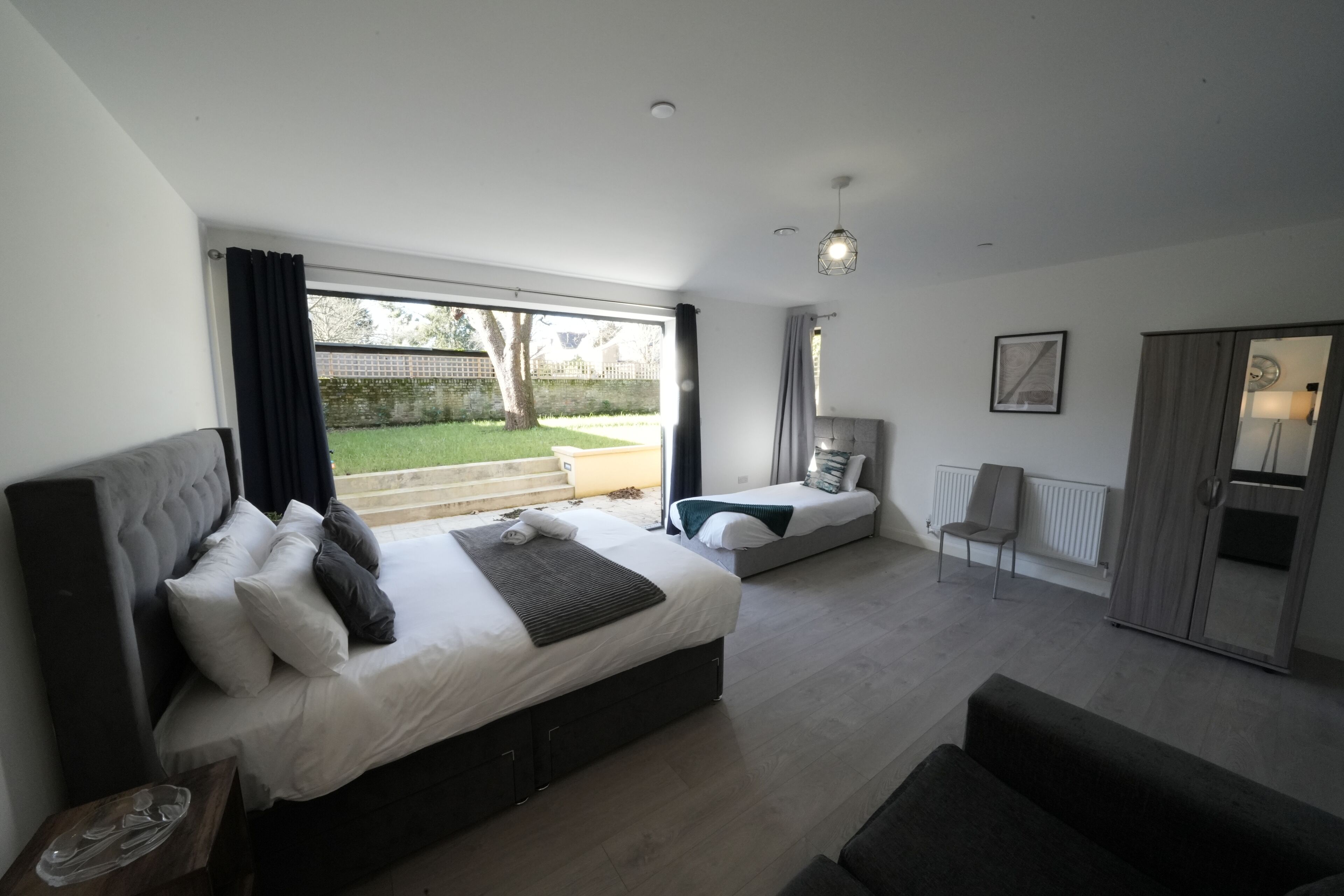 A modern bedroom with a large bed, a smaller bed, a sofa, a chair, and a view of the garden.