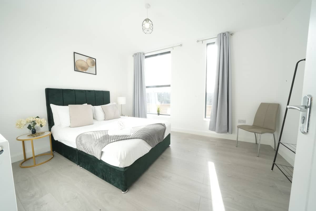 A modern bedroom with a large bed, a side table with flowers, a chair, and a window with curtains.