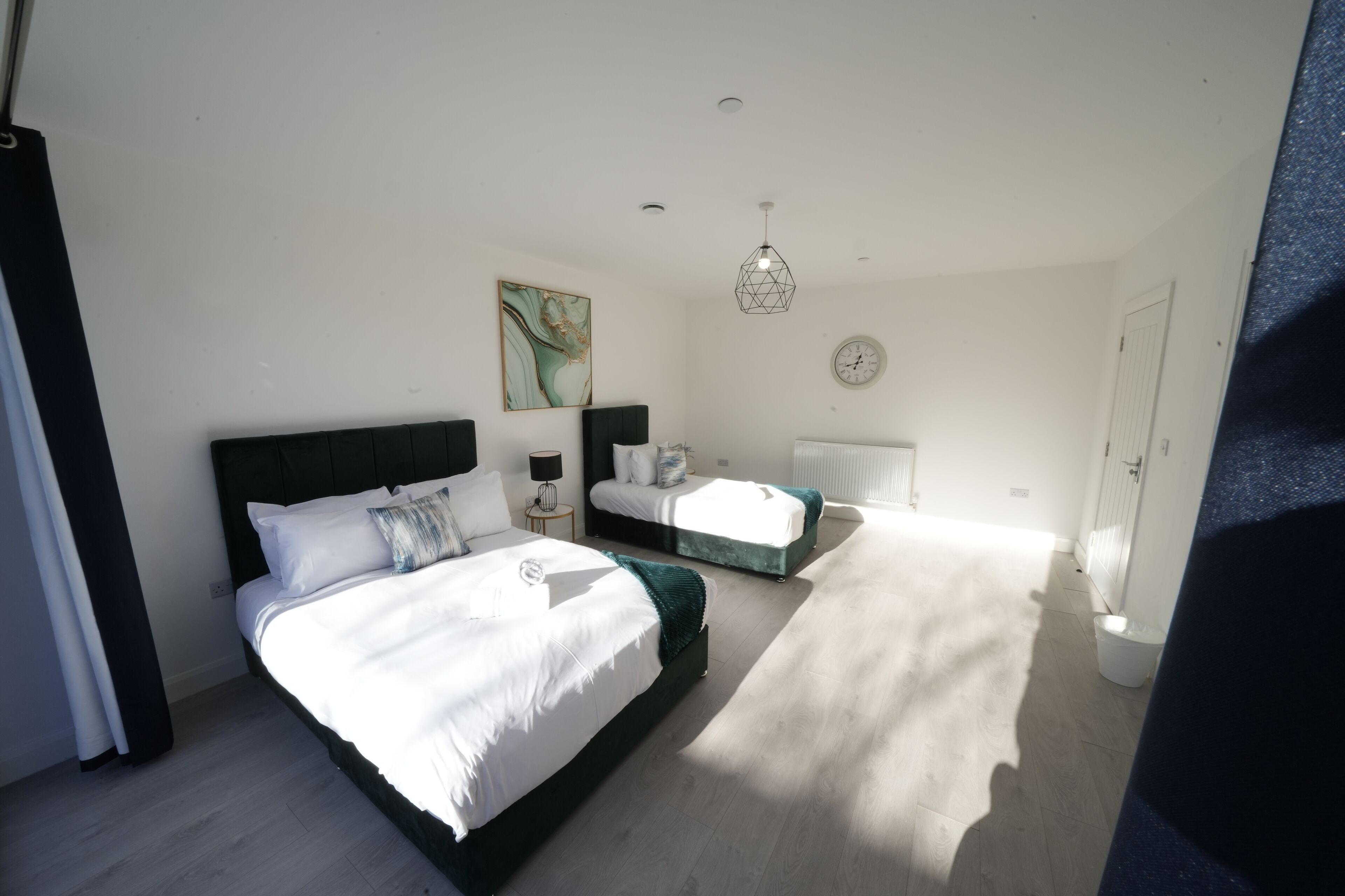 A modern bedroom with a large bed, a smaller bed, a nightstand, a lamp, a wall clock, and a ceiling light.