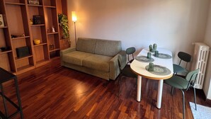 A living room with a wooden floor, a green sofa, a dining table with chairs, and a bookshelf.