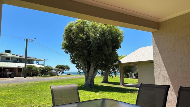 A covered outdoor seating area with a table and chairs, a large tree, and a view of the ocean. - Pelican Shore Villa 20 - Kalbarri WA (Kalbarri)