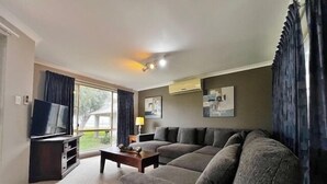 A living room with a sofa, a coffee table, a TV, and a window with curtains. - Pelican Shore Villa 20 - Kalbarri WA (Kalbarri)