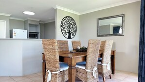 A dining area with a wooden table and wicker chairs, a wall-mounted tree decal, and a kitchen in the background. - Pelican Shore Villa 20 - Kalbarri WA (Kalbarri)