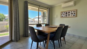Un coin repas avec une table en bois et quatre chaises, de grandes fenêtres avec des rideaux et un appareil de climatisation fixé au mur.