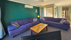 A living room with a green wall, two purple sofas, a wooden coffee table, a TV, and an air conditioning unit. - Pelican Shore Villa 22 - Kalbarri WA (Kalbarri)
