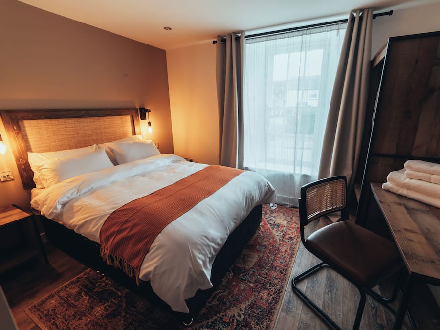 A hotel room with a large bed, a desk with a chair, a window with curtains, and a wooden headboard.