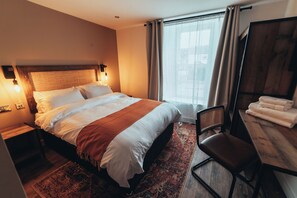 Hotellihuone, jossa on suuri sänky, työpöytä tuolilla, ikkuna verhoilla ja puinen pääty.