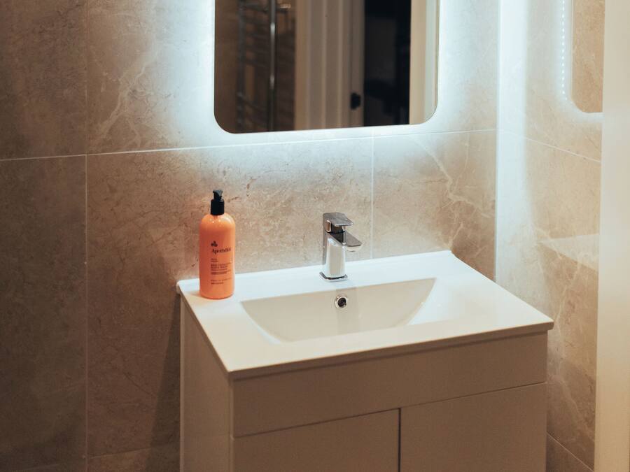 Un baño con lavamanos blanco, un espejo y una botella de champú.