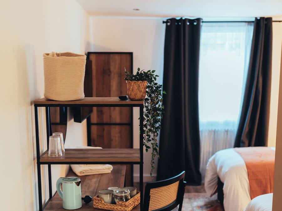 Una habitación de hotel con suelo de madera, una cama con manta naranja, una mesita con una planta, una silla y una estantería con varios objetos.