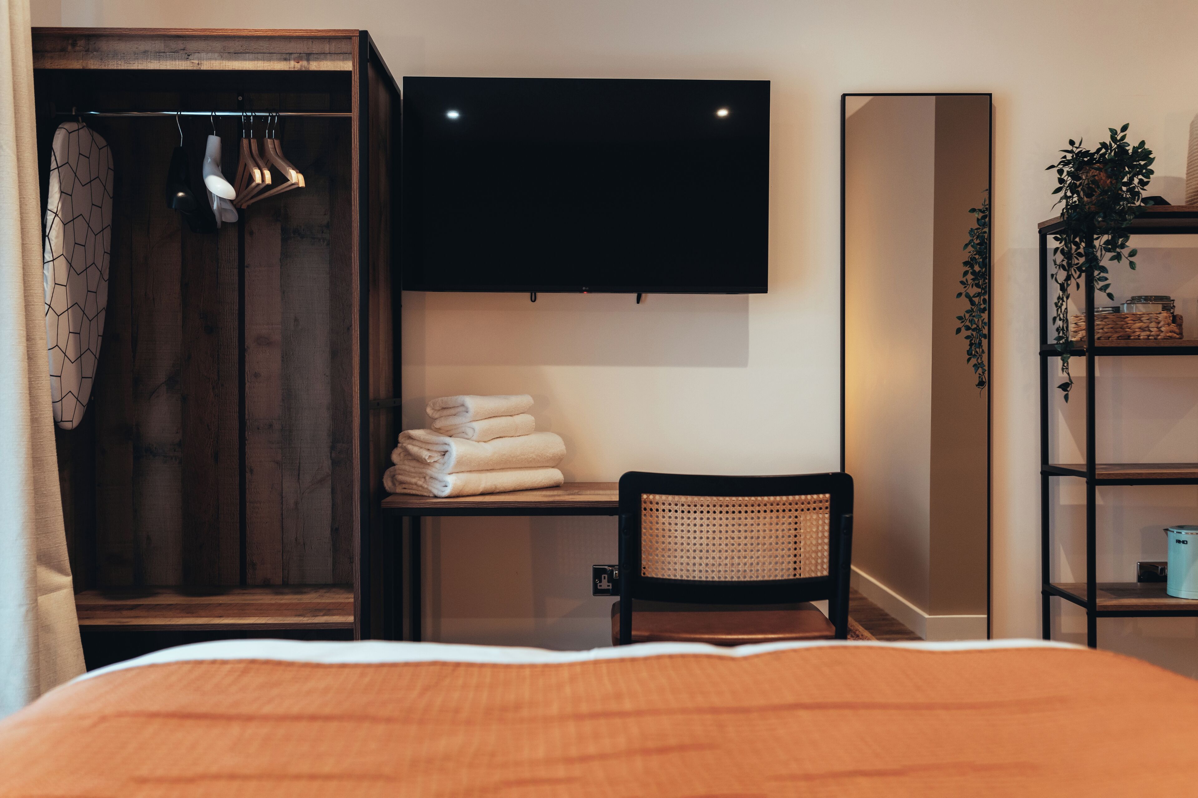 Quarto com cama, guarda-roupa de madeira, uma cadeira, escrivaninha, televisão e prateleira com plantas.