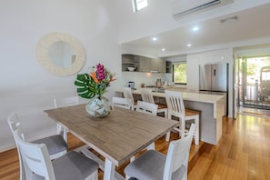 Una cocina moderna con armarios blancos, un refrigerador de acero inoxidable y un comedor con mesa de madera y sillas blancas.