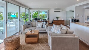 A modern living room with a sofa, ottoman, and coffee table, a dining area with a table and chairs, and a kitchen area with a counter and appliances. - Oasis 21, Oasis 21 on Hamilton island (Hamilton Island)